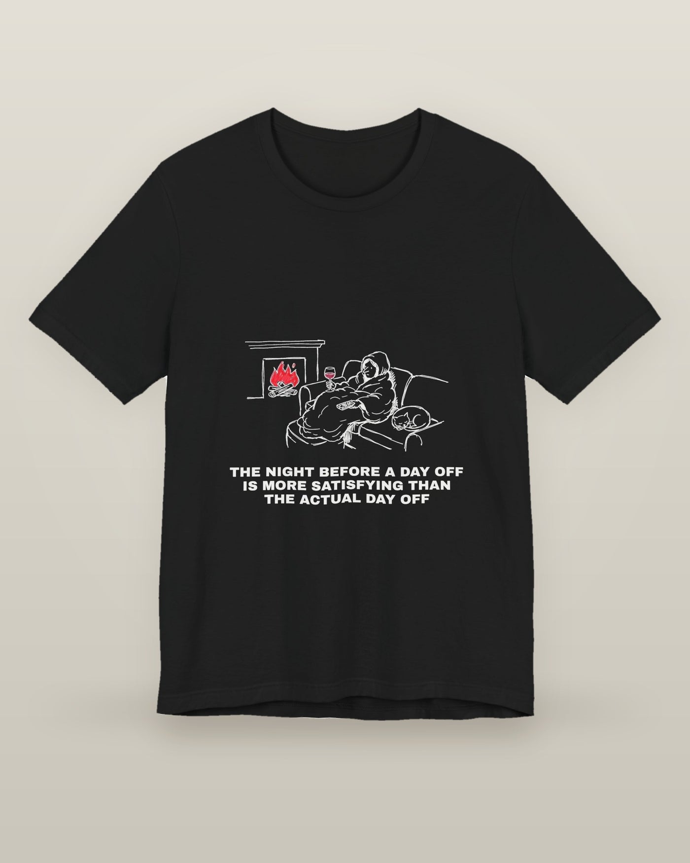 The night before a day off is more satisfying than the actual day off — a hooded figure under a blanket with wine, a remote, a sleeping cat and a lit fire, printed on a black cotton t-shirt, front view