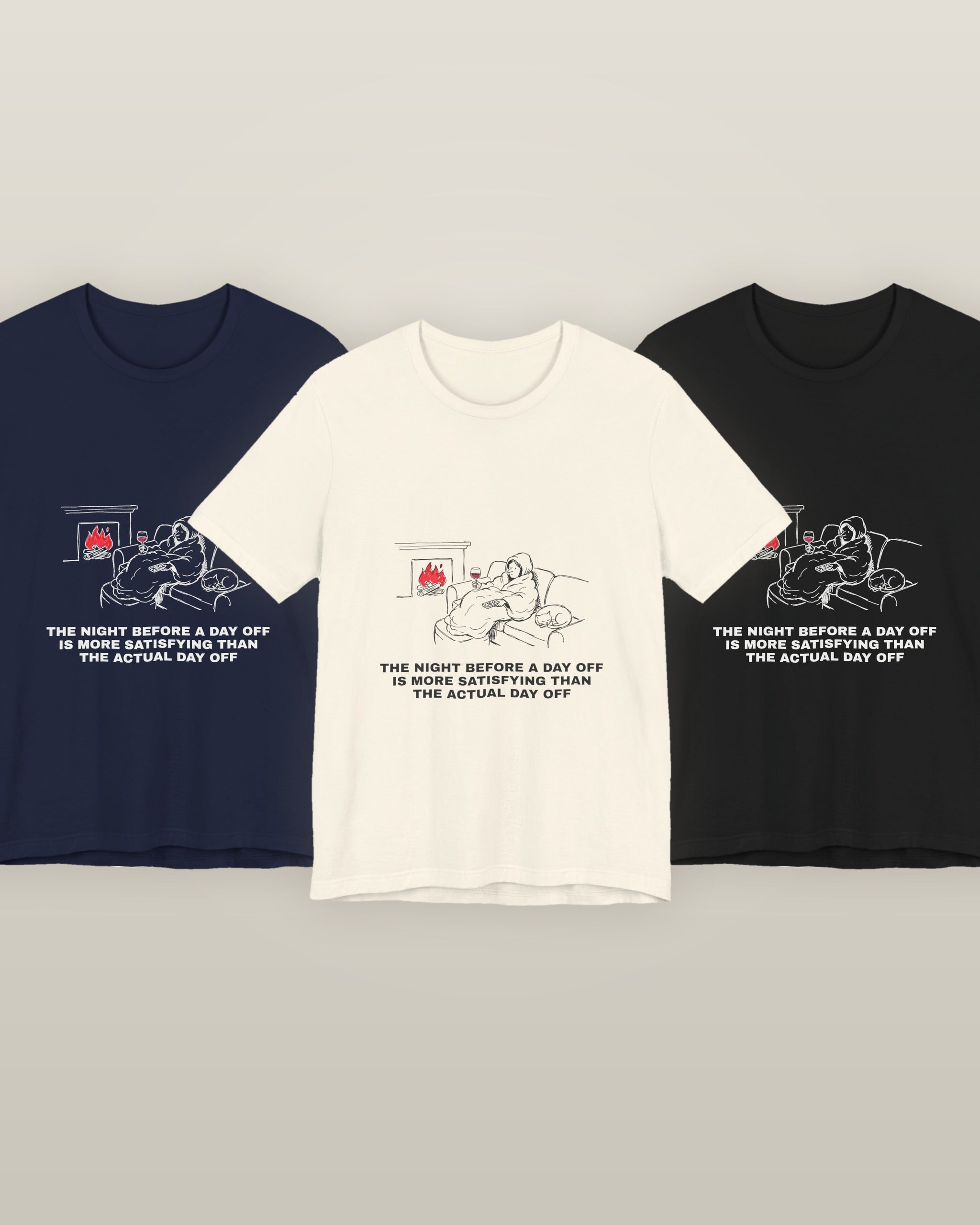 The night before a day off is more satisfying than the actual day off — a hooded figure under a blanket with wine, a remote, a sleeping cat and a lit fire, printed on a natural cream, navy and black cotton t-shirt, all three colourways together