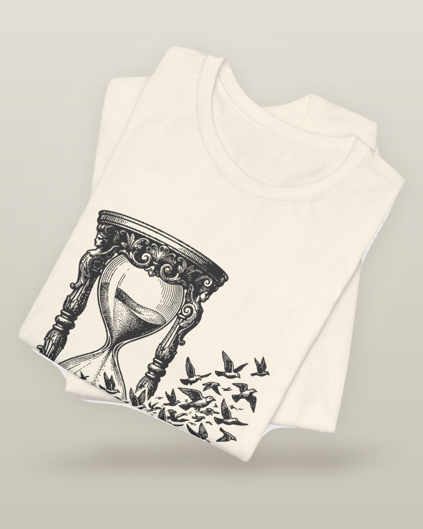 The time I gave away — an hourglass whose falling sand becomes a flock of birds, printed on a natural cream cotton t-shirt, folded