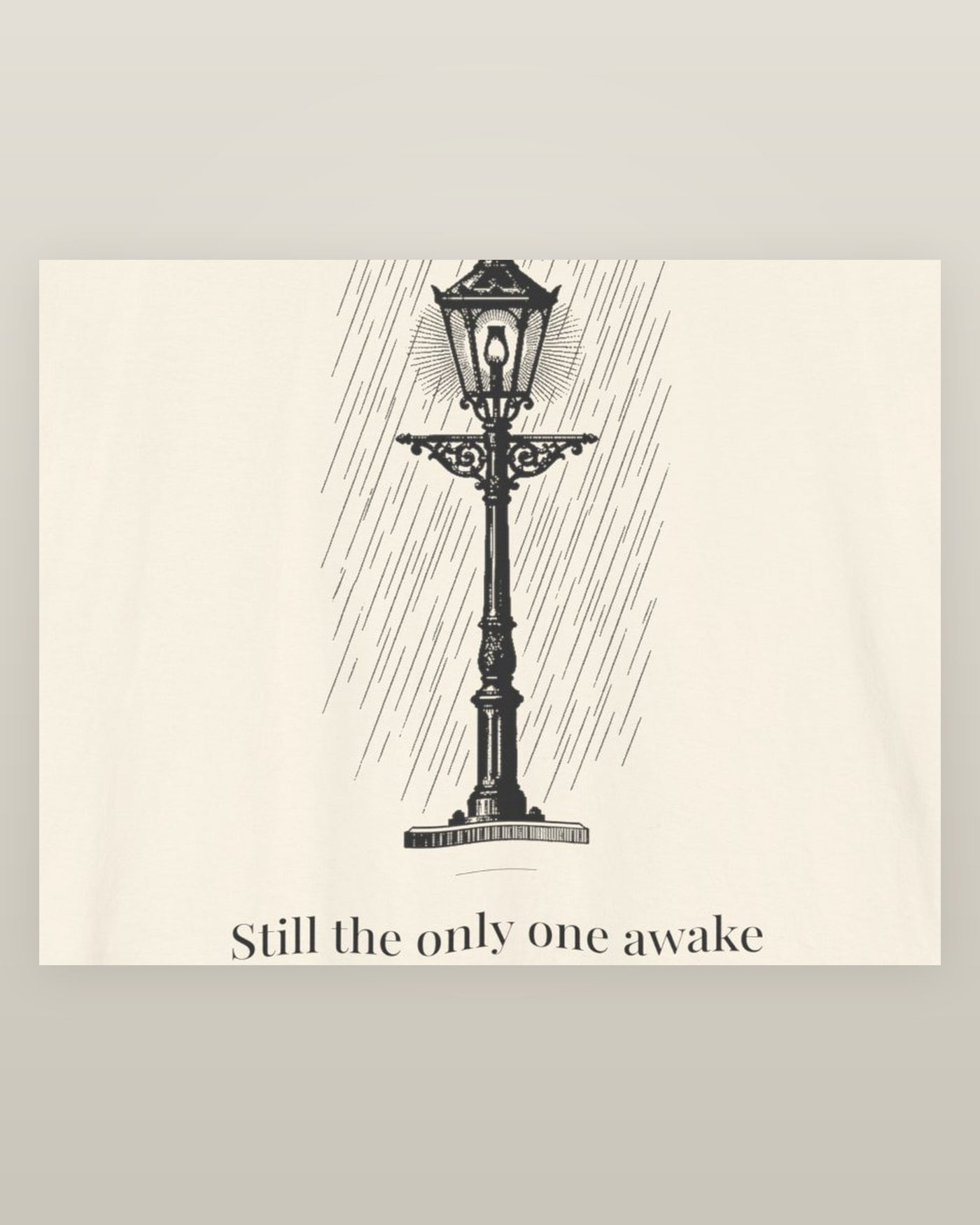Still the only one awake — a cast-iron street lamp lit in fine rain, printed on a natural cream cotton t-shirt, close detail of the print