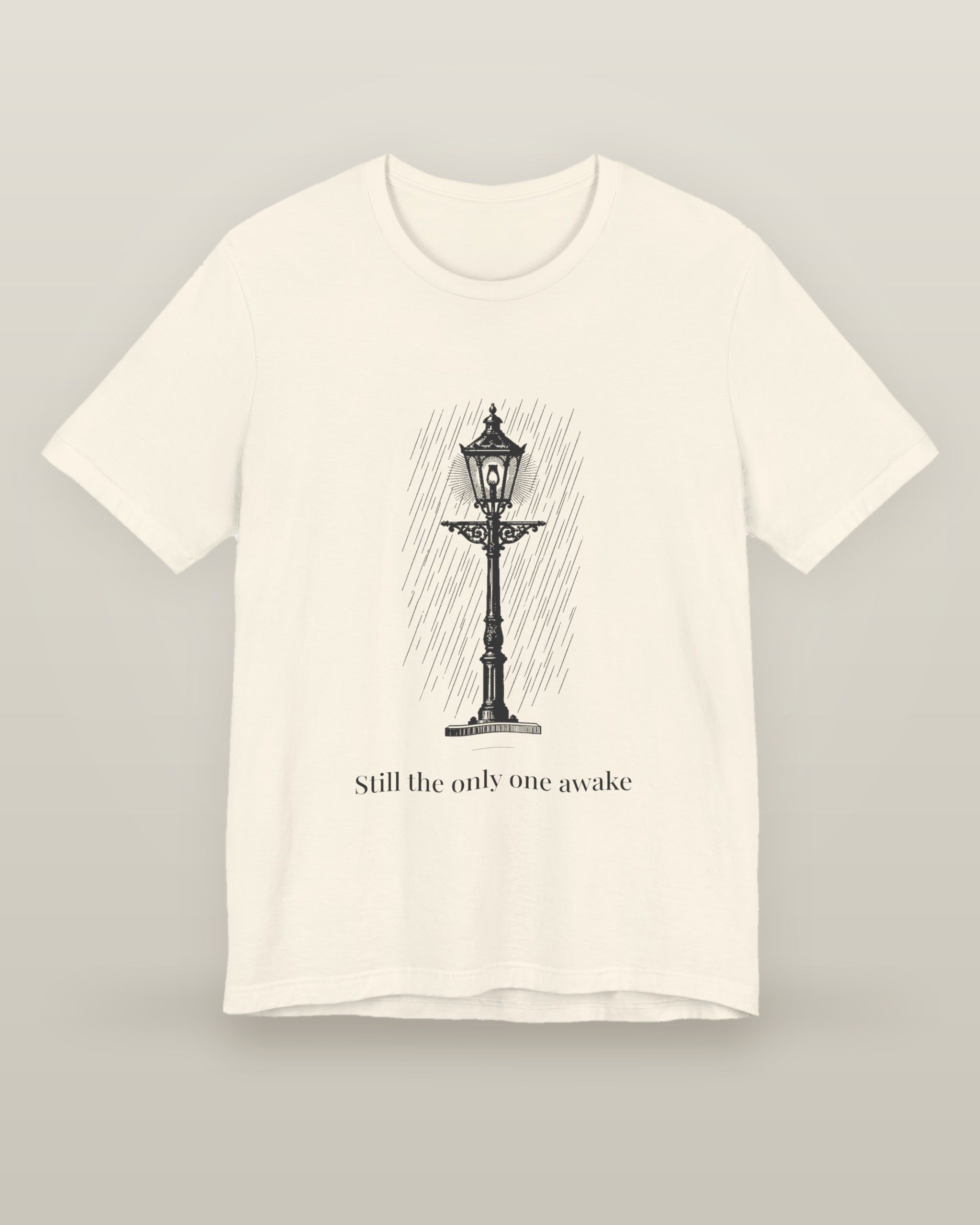 Still the only one awake — a cast-iron street lamp lit in fine rain, printed on a natural cream cotton t-shirt, front view