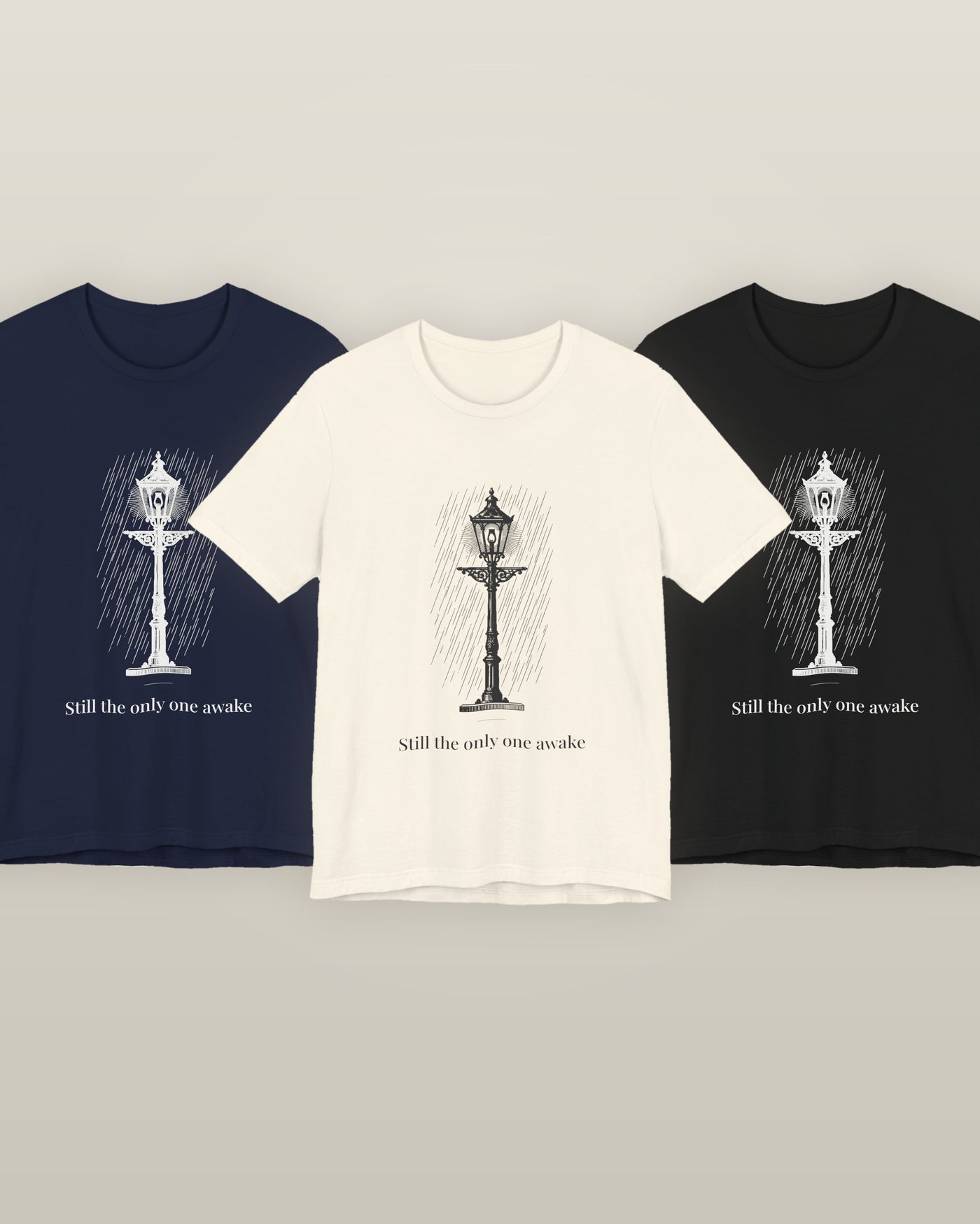 Still the only one awake — a cast-iron street lamp lit in fine rain, printed on a natural cream, navy and black cotton t-shirt, all three colourways together