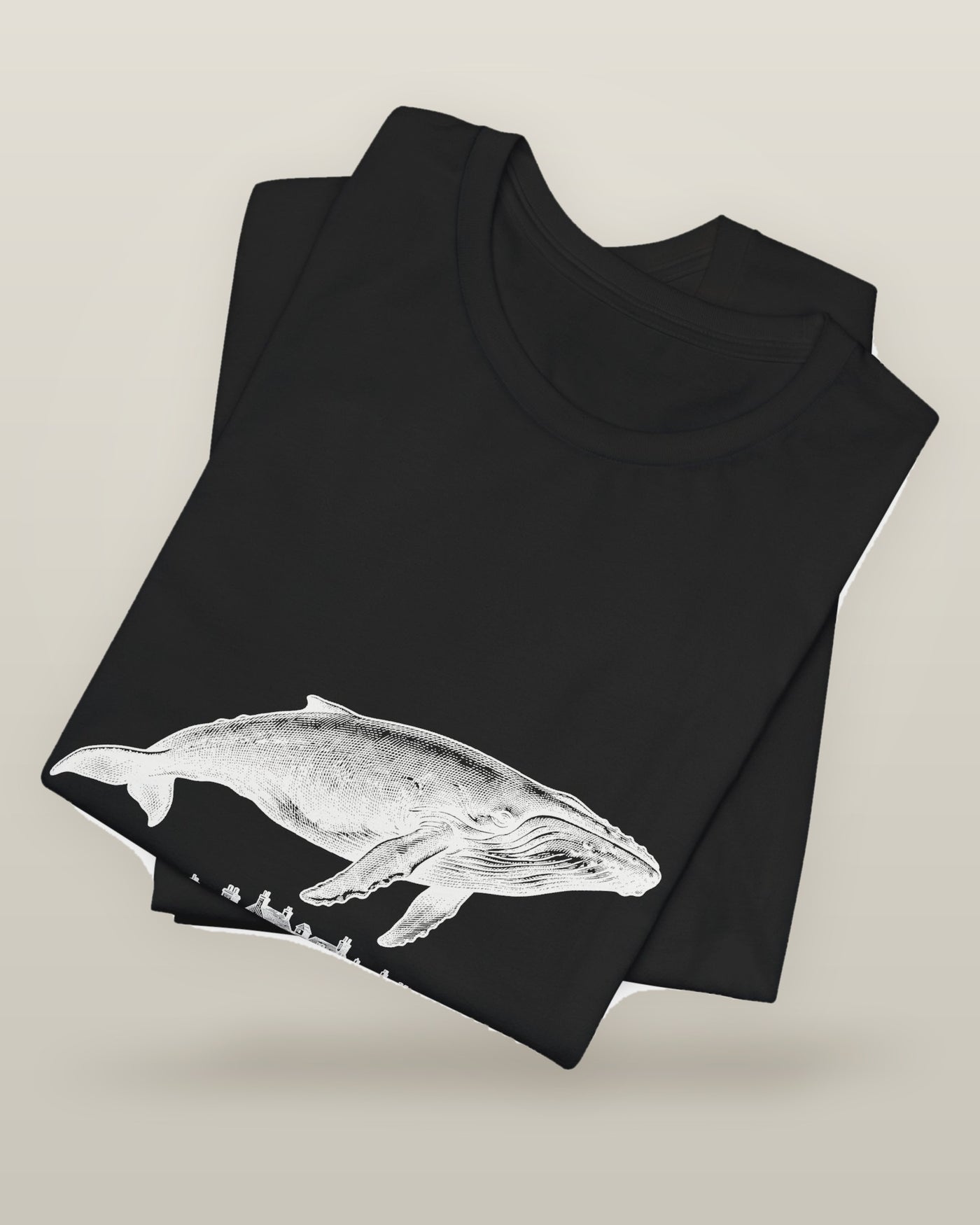 I outgrew the water — a humpback whale floating above rooftops, printed on a black cotton t-shirt, folded