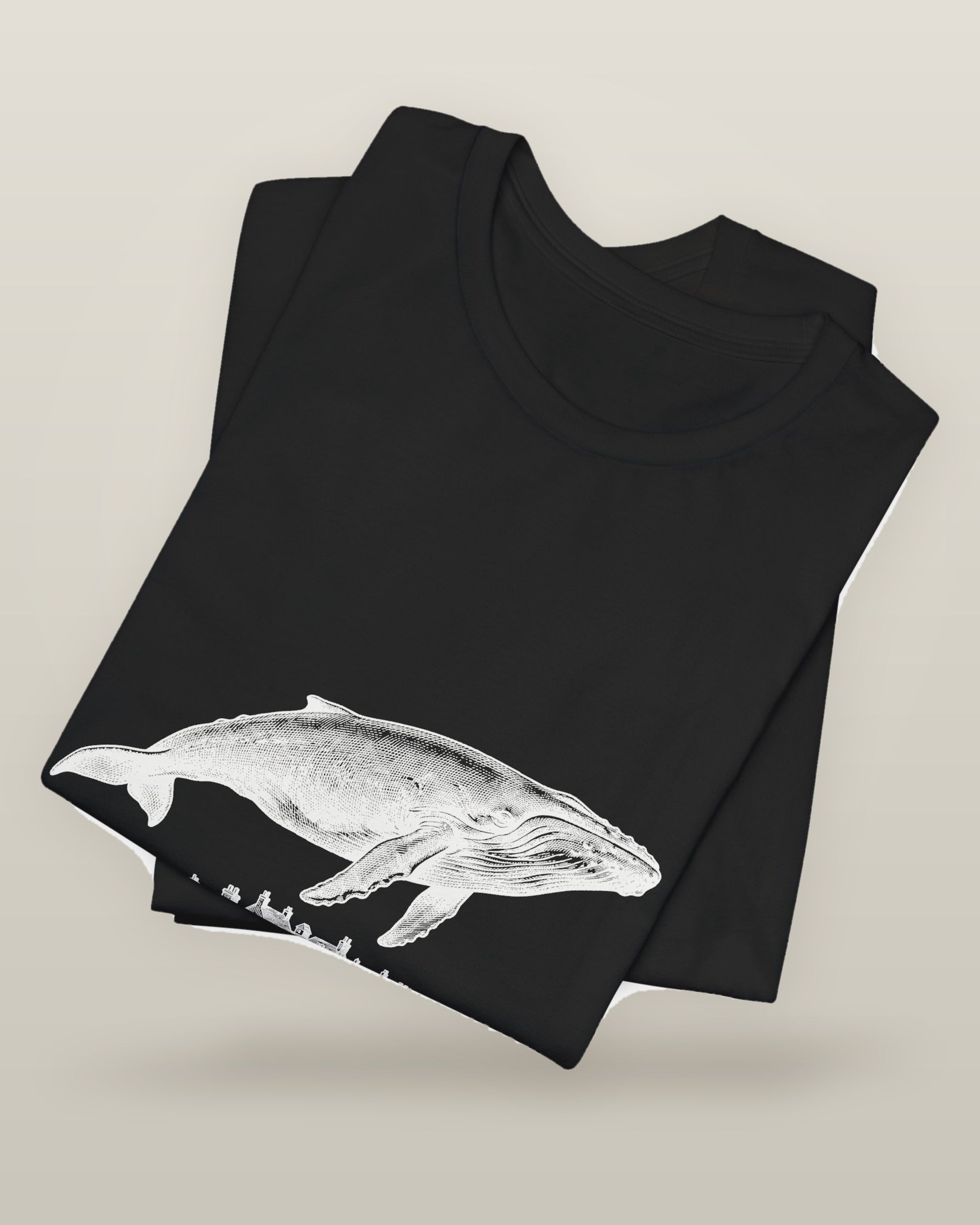 I outgrew the water — a humpback whale floating above rooftops, printed on a black cotton t-shirt, folded