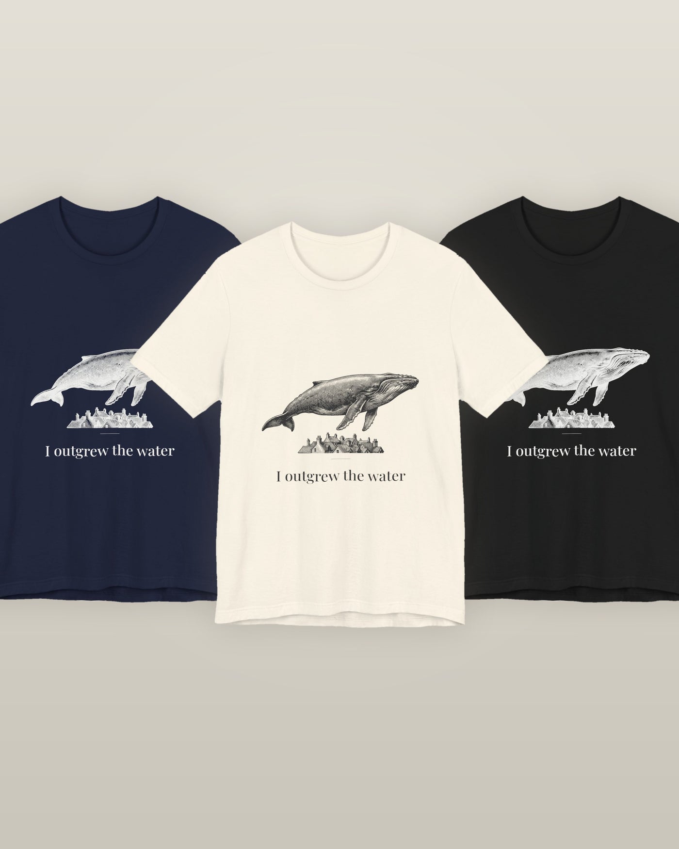 I outgrew the water — a humpback whale floating above rooftops, printed on a natural cream, navy and black cotton t-shirt, all three colourways together