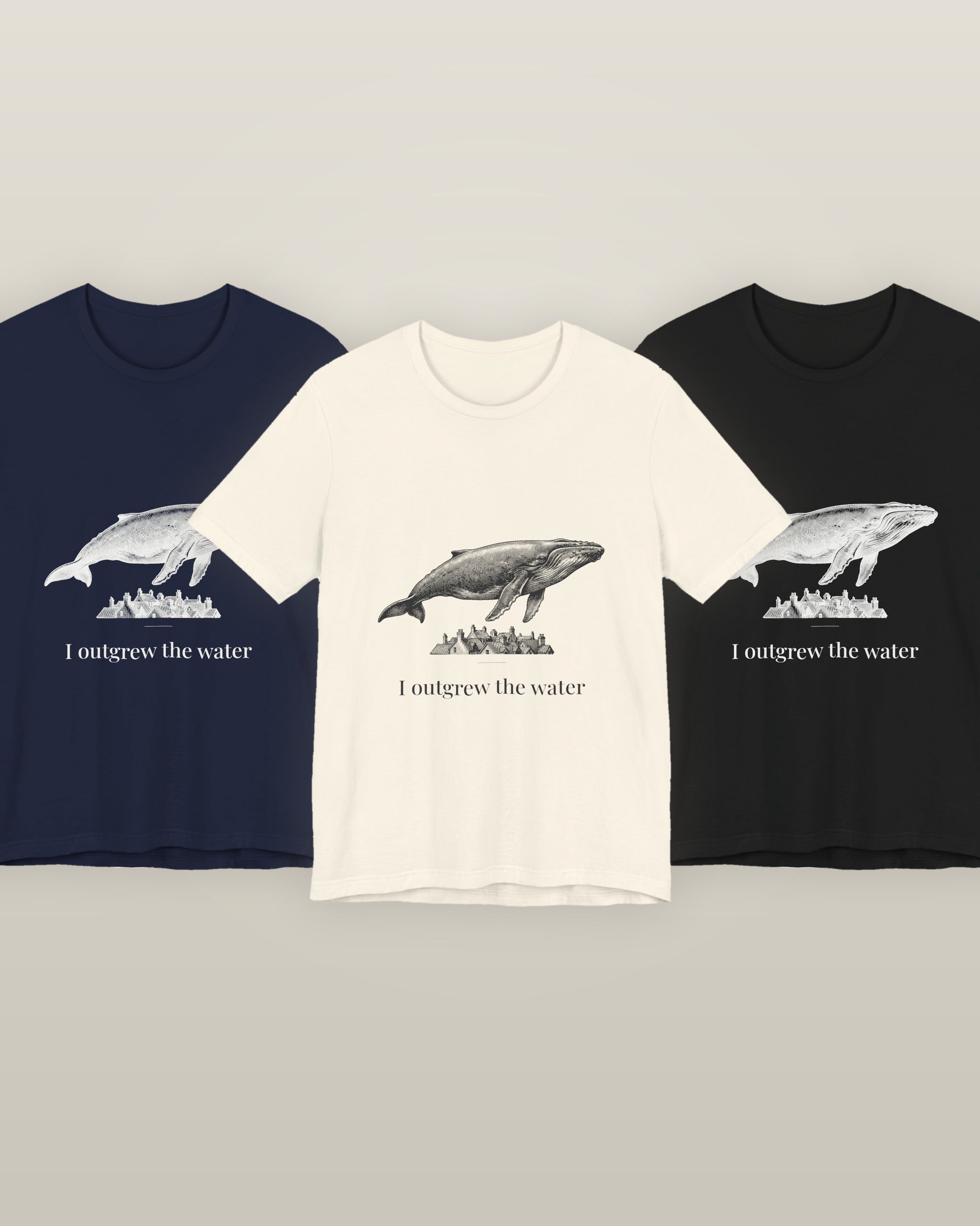 I outgrew the water — a humpback whale floating above rooftops, printed on a natural cream, navy and black cotton t-shirt, all three colourways together